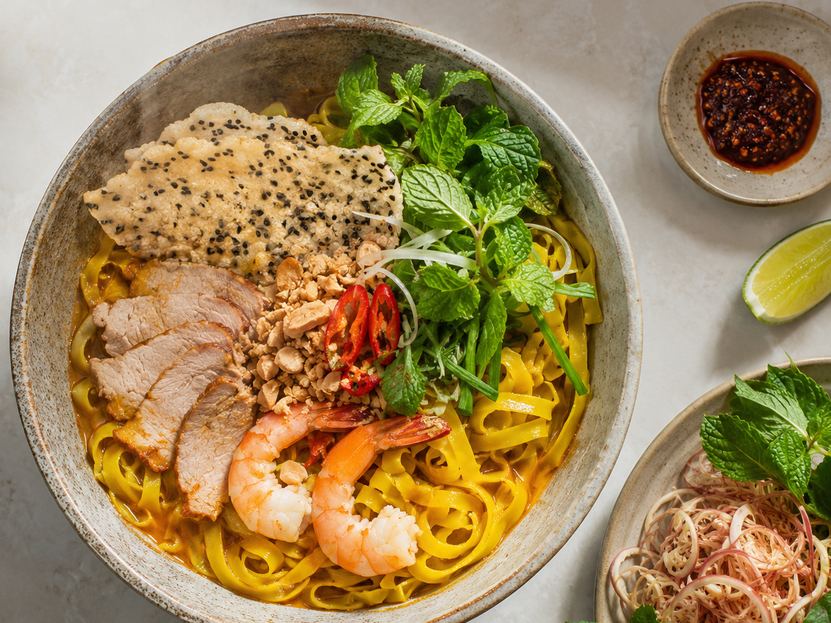 A bowl of mì Quảng with pork, shrimp, peanuts, herbs and a sesame rice cracker